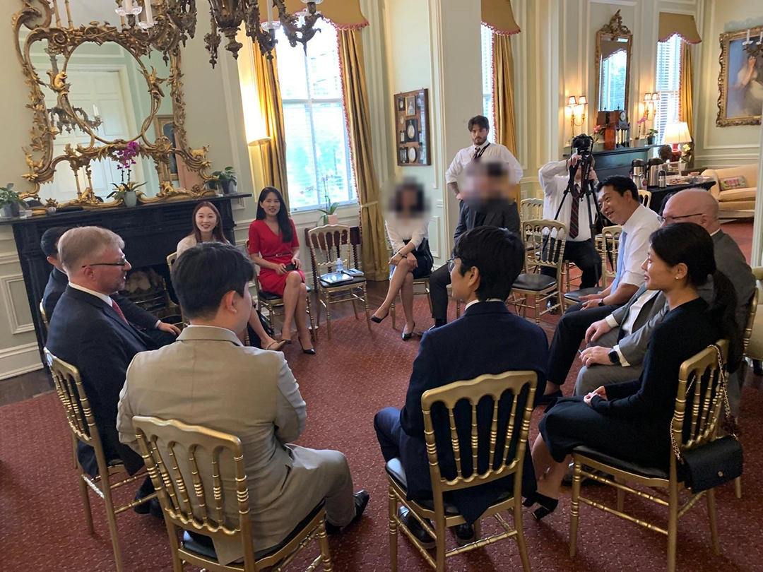 A group of people in formal attire sit in a circle in a well-decorated room, engaged in conversation. Photographers and recording equipment are present.
