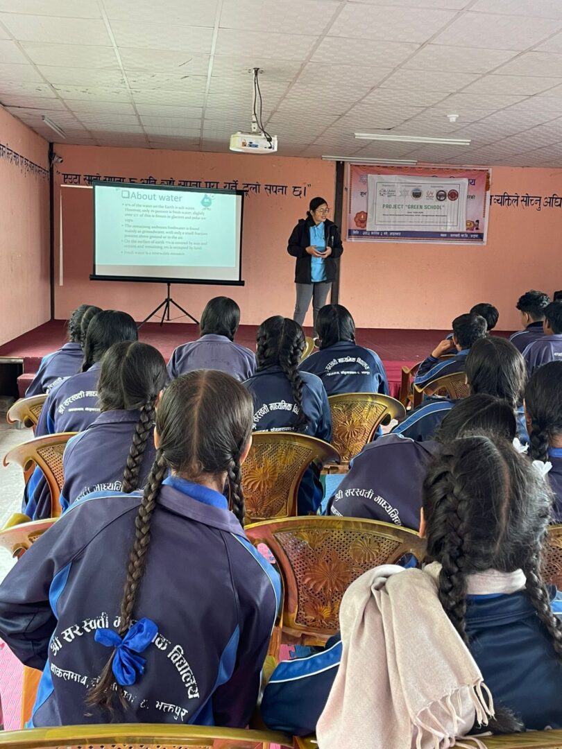 A woman from GPF Nepal gives a presentation about water to students in uniform, highlighting Knowledge and Service. A projector screen displays information at the front of the classroom.