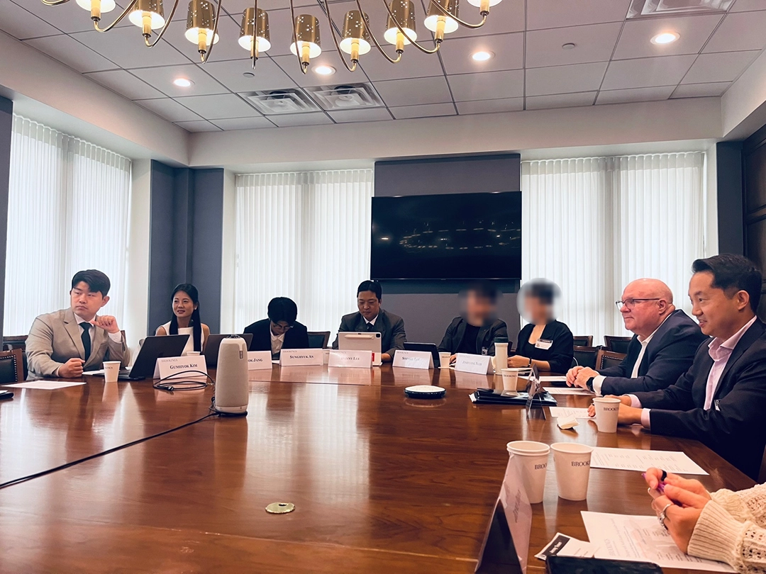A group of people in business attire sit around a large conference table with laptops, papers, and nameplates in a meeting room.