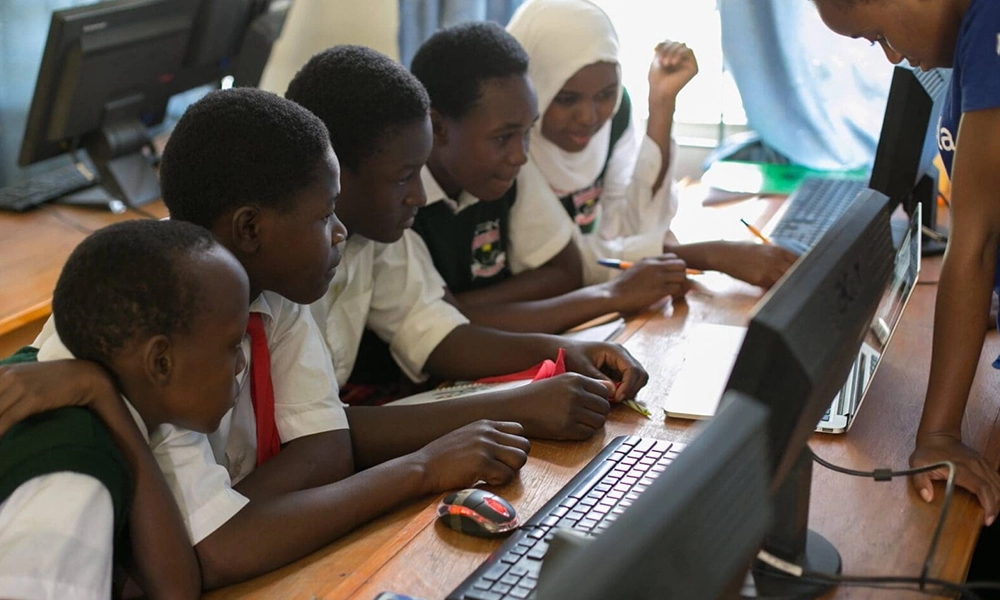 Five students in school uniforms sit at a desk, working together on a computer, while an adult assists them in a classroom with other computers visible.