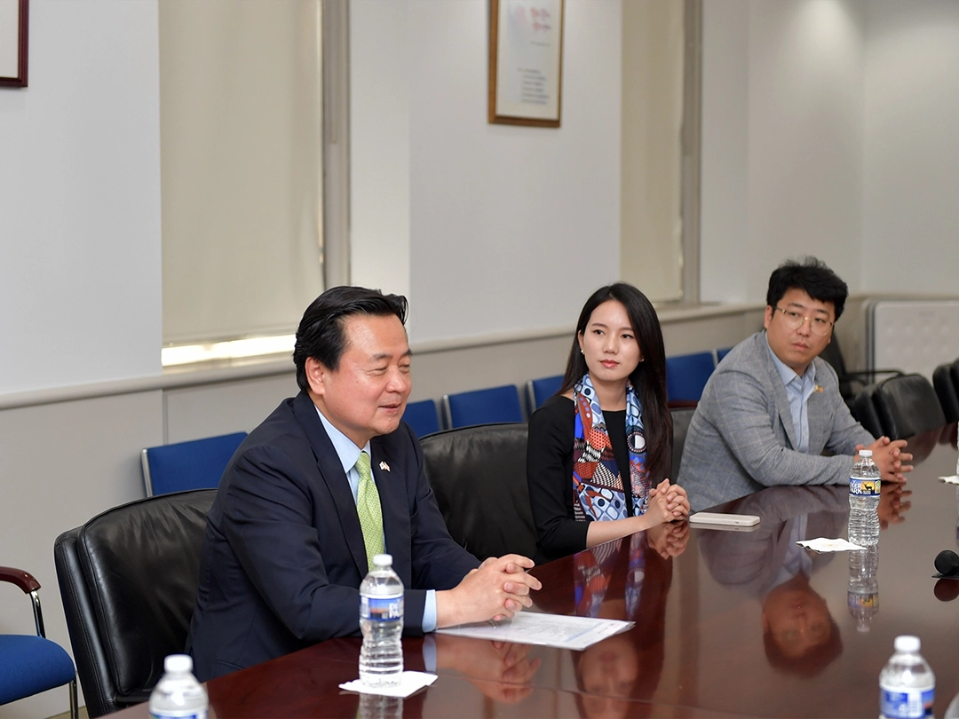 Three people in business attire sit at a conference table with bottled water and papers, engaged in discussion in a meeting room.