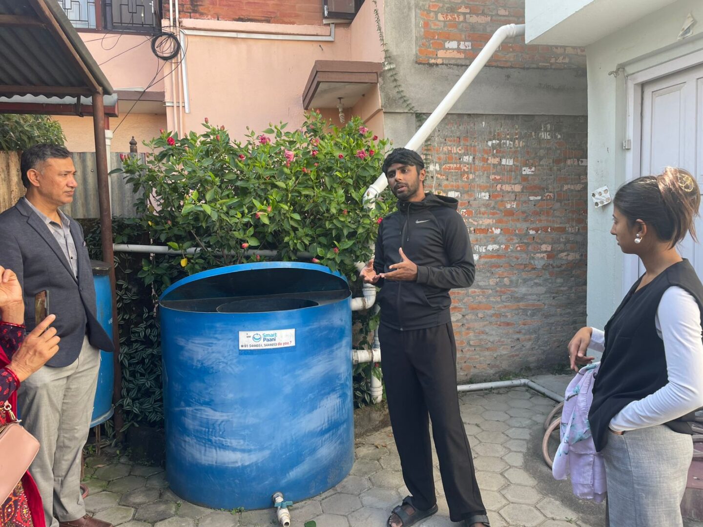 A man in black speaks to three people near a large blue water tank in an outdoor area with brick walls and plants, highlighting GPF Nepal's commitment to Sustainable Communities.