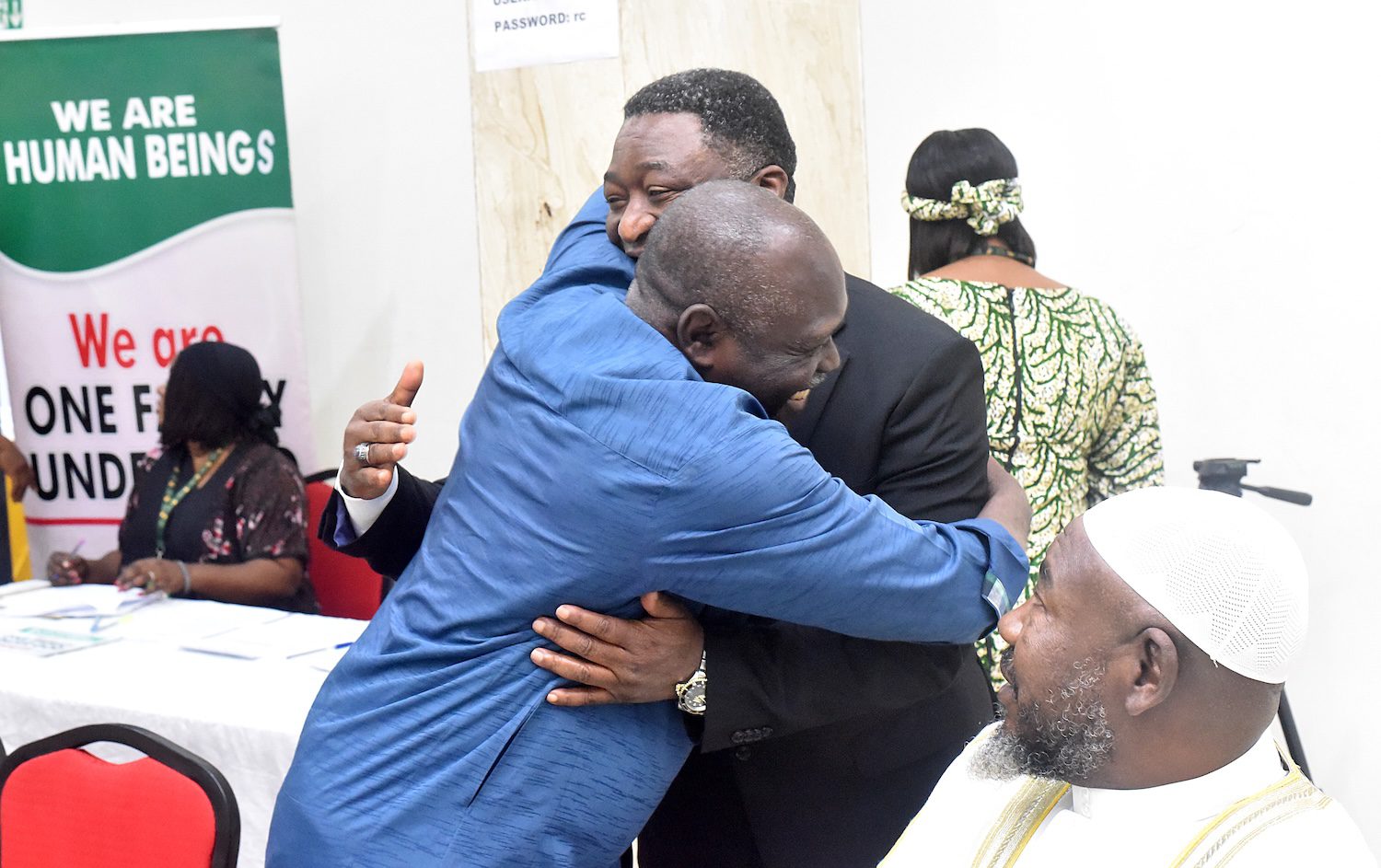 Two faith leaders in suits embrace each other in a hug, while another man in white sits nearby. A sign in the background reads,