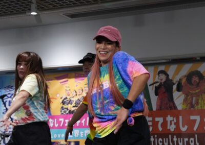A group of people in colorful tie-dye shirts and casual hats are dancing indoors, smiling, at a Multicultural Family Festival, with festival posters for 2025 visible in the background.