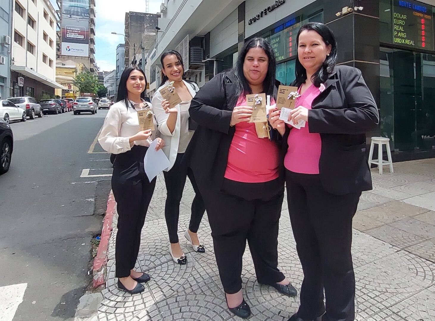 Four women in business attire stand on a city sidewalk in Paraguay, holding brown paper bags and smiling at the camera, embodying women empowerment. Buildings and parked cars are visible in the background.