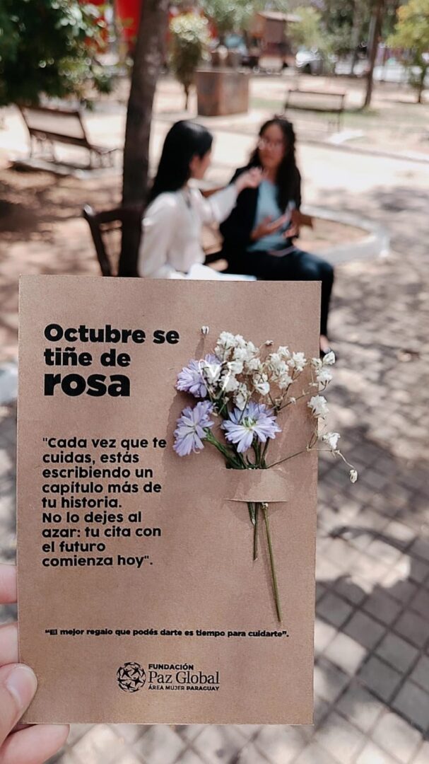 A hand holds a card with flowers attached, featuring Spanish text about self-care for Pink October, while two women in Paraguay sit and talk on a bench in the blurred park background.