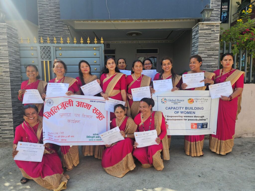 A group of women in matching red sarees stand outside, holding certificates and banners about a capacity-building program for women focused on strengthening families and peacebuilding.
