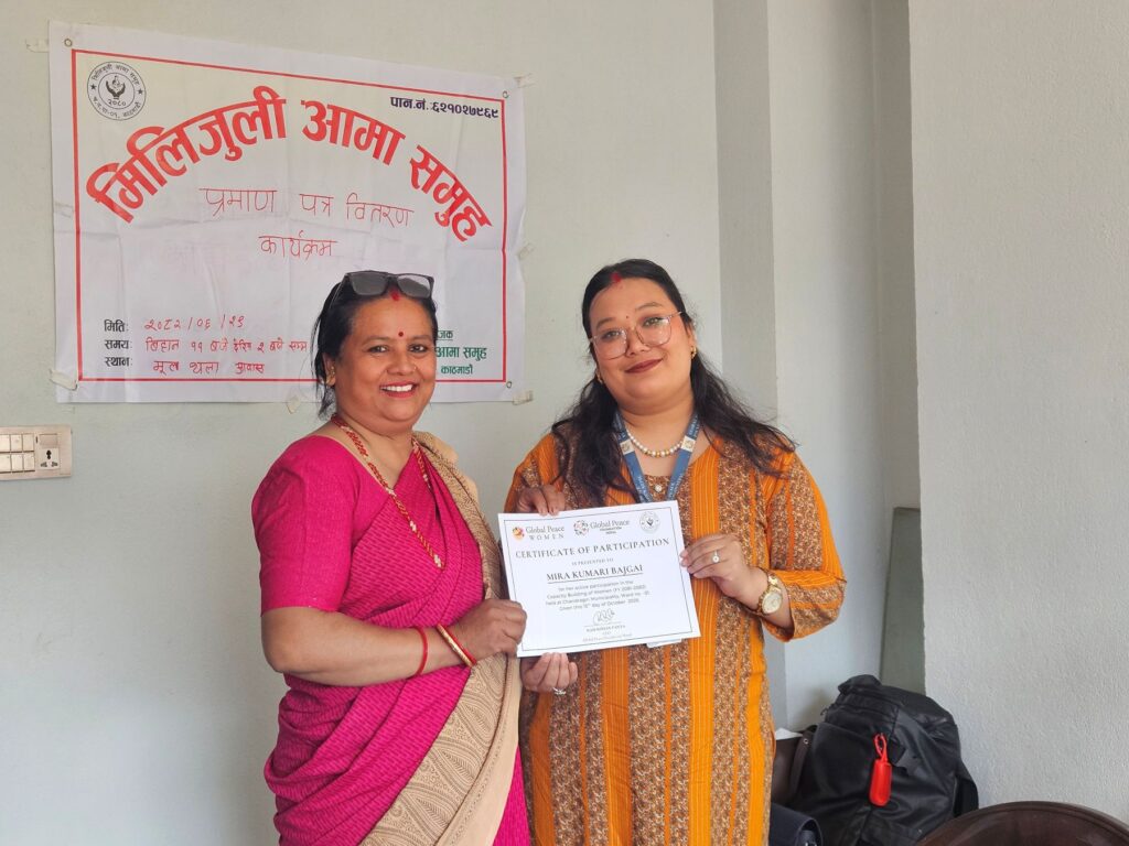 Two women stand indoors, one in pink and one in yellow, smiling as the woman in yellow holds a certificate of participation. A banner in Nepali about strengthening families is displayed on the wall behind them.