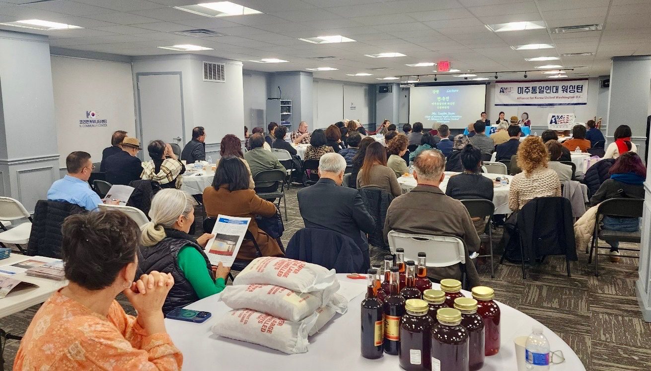 A diverse group of people, including North Korean escapees, attends an indoor seminar; pamphlets, bottled drinks, and bags of rice are placed on a table in the foreground.