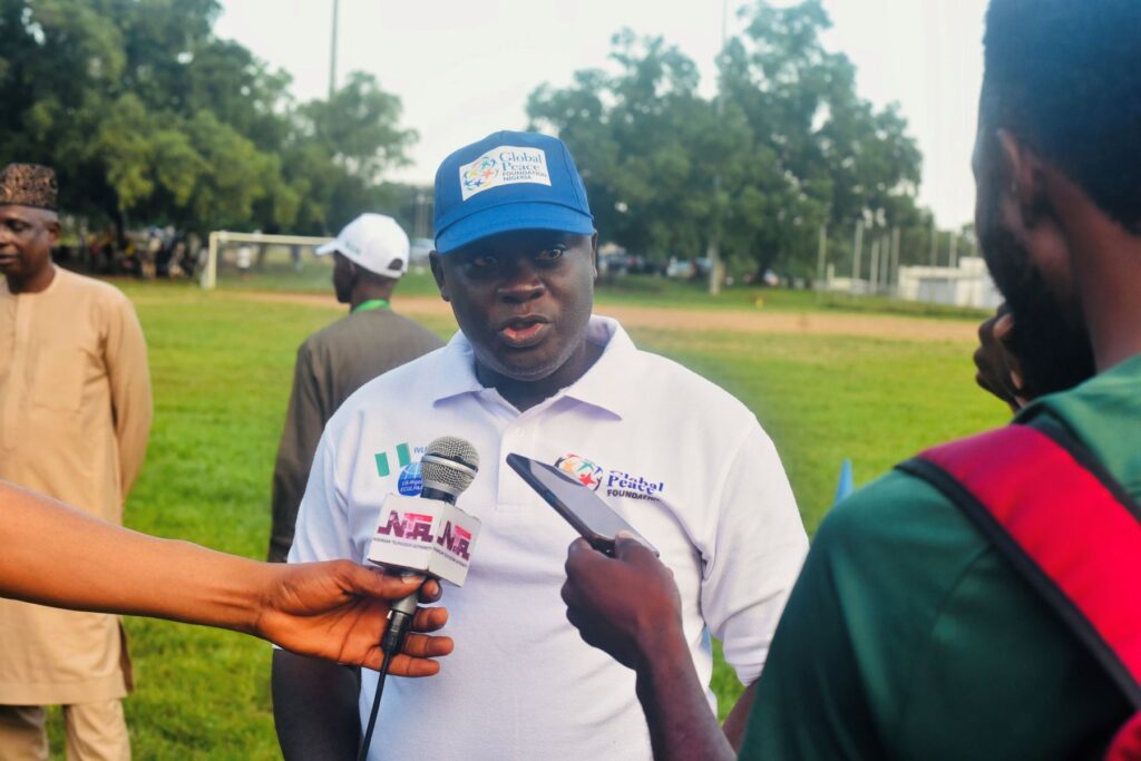 A man wearing a white polo shirt and a blue cap speaks to reporters holding microphones outdoors on a grassy field, highlighting the role of sports in promoting unity and peace.