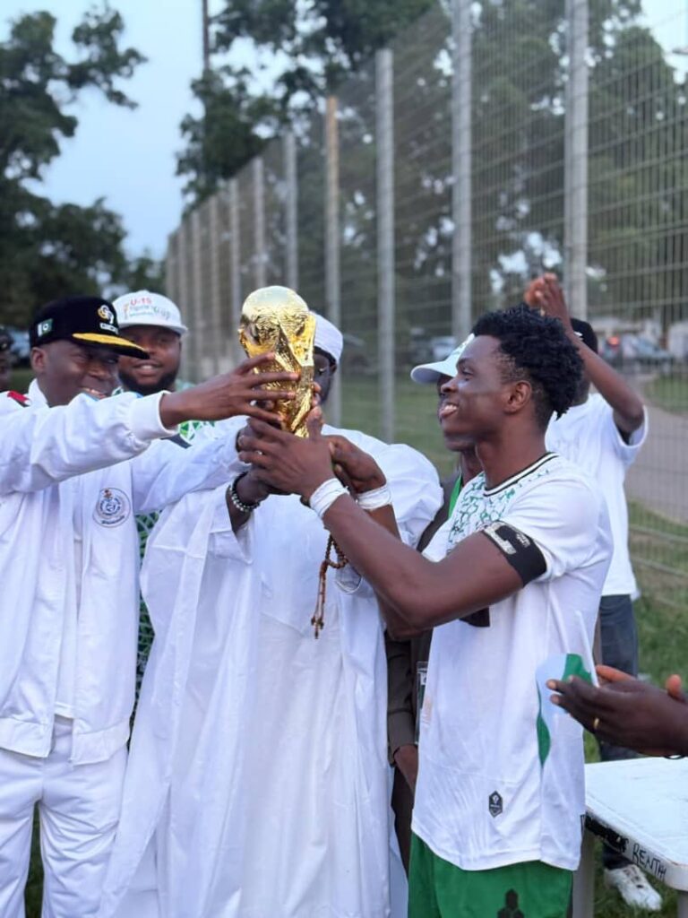 A young male soccer player in a white jersey receives a golden trophy from officials during an outdoor ceremony, celebrating unity in sports as people clap and smile in the background.