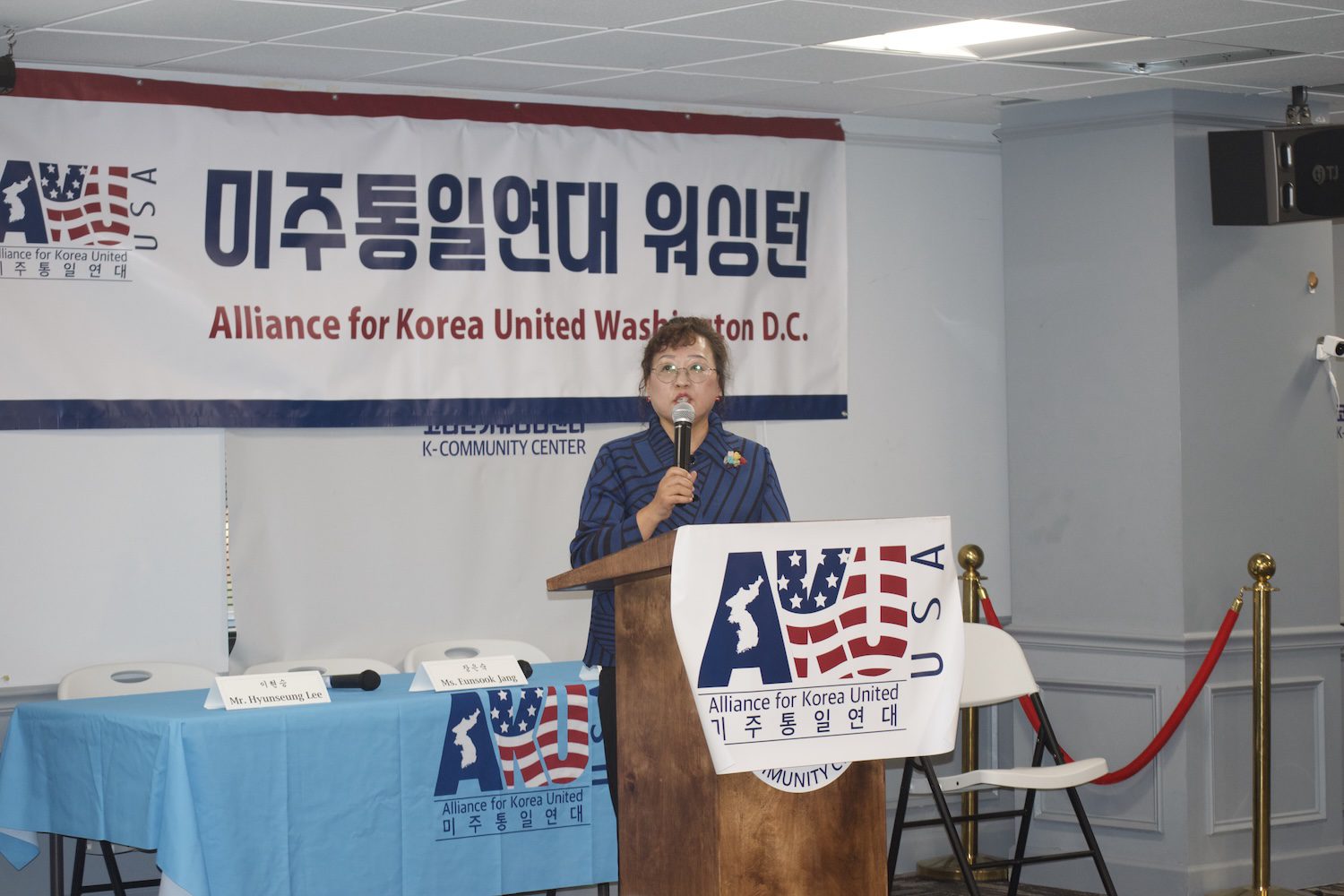A woman stands at a podium speaking during an AKU Washington talk concert in front of a banner that reads