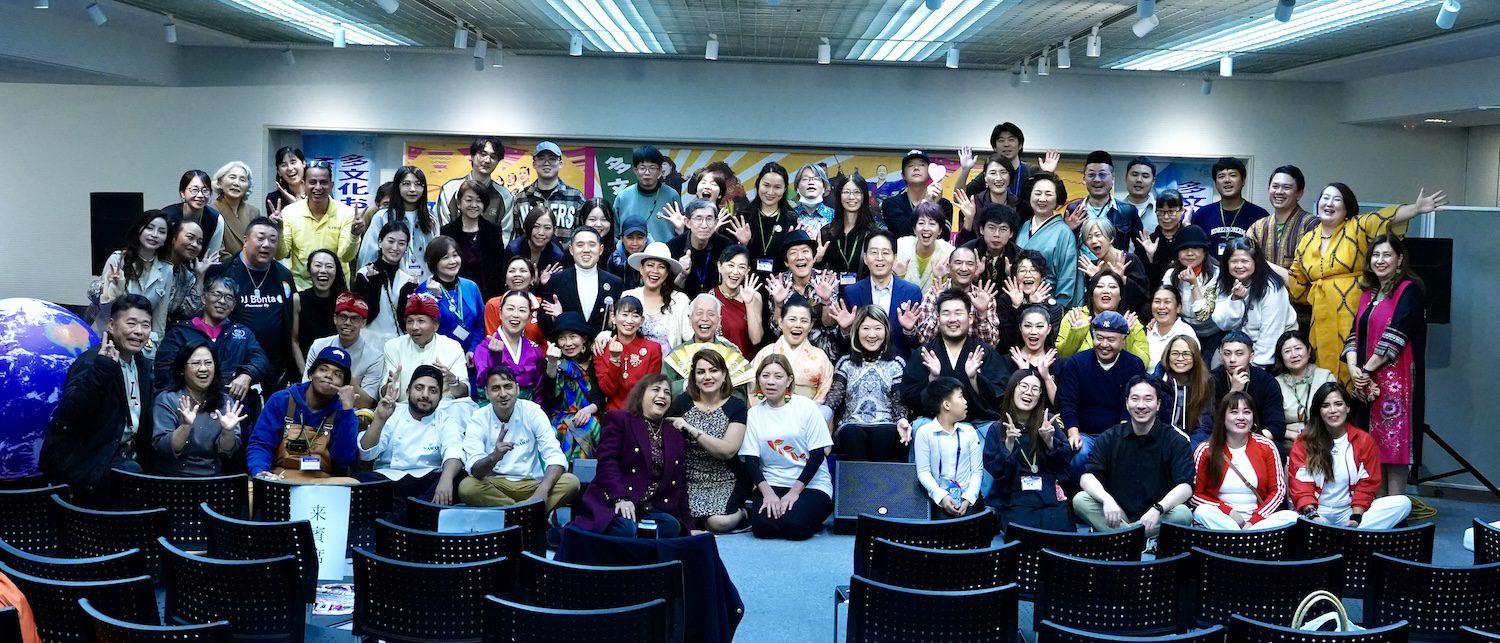 A large group of people pose together indoors for a group photo at the 2025 Multicultural Festival, smiling and waving at the camera; rows of empty chairs are visible in the foreground.