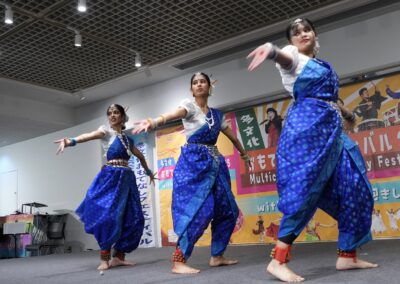 Three women in traditional blue attire perform a classical Indian dance on stage at the 2025 One Family Festival, with colorful banners and posters in the background.
