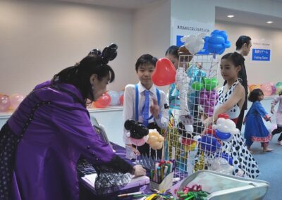 At the 2025 One Family Festival, a woman in costume delights children in dressy clothes by making balloon animals at a table filled with colorful balloons and markers.
