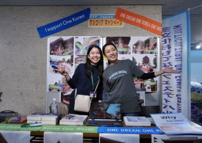 Two women stand and smile at an informational booth promoting the "One Dream One Korea One World" campaign and the upcoming 2025 One Family Festival, with posters, banners, and pamphlets displayed around them.