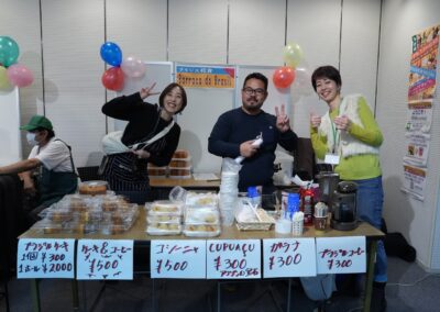 Three people stand behind a table at a market stall selling Brazilian food and drinks at the 2025 One Family Festival, part of a lively multicultural festival. Signs in Japanese display prices, and colorful balloons decorate the background.