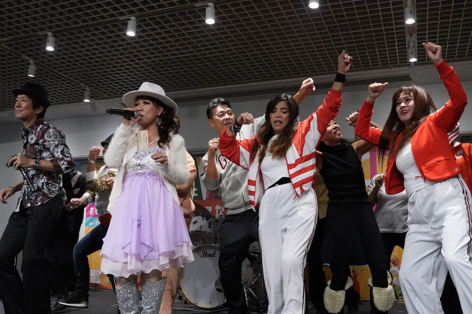 A group of people dance enthusiastically on stage at a Family Festival while a woman in a white hat and lavender dress sings into a microphone. A drum set is visible in the background, adding to the lively, multicultural atmosphere.