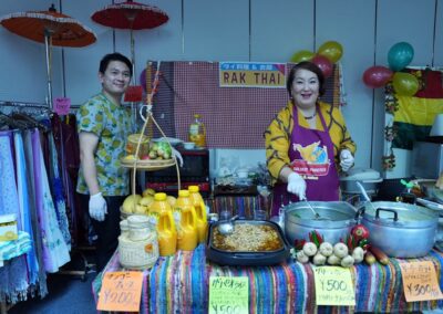 Two people stand behind a table with Thai food, drinks, and produce at the "RAK THAI" market stall during the 2025 Multicultural Festival. Colorful fabrics and decorations surround them, showcasing vibrant cultural diversity.