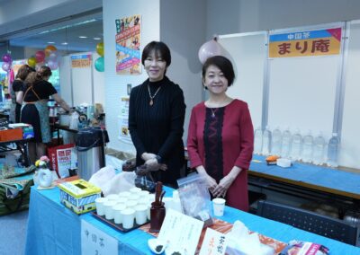 Two women stand behind a table with cups and tea supplies at a Multicultural Family Festival in 2025, smiling at the camera. Signs and balloons decorate the festive indoor area.