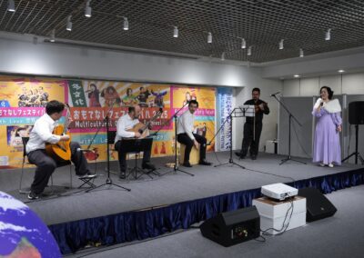Five performers are on stage at the 2025 Multicultural Festival: three playing guitars, one on violin, and one singing into a microphone, with colorful banners hanging behind them.