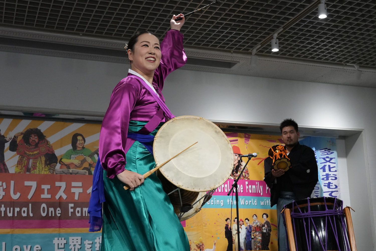 At a vibrant 2025 Family Festival, a woman in traditional Korean attire plays a janggu drum on stage, smiling, as two musicians accompany her on drums in the background, creating a joyful multicultural celebration.