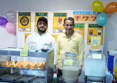 Two men stand behind a food stall at the 2025 One Family Festival, selling samosas, ice tea, and lassi. Colorful balloons decorate the stand, and menu signs with prices are displayed on the wall behind them.