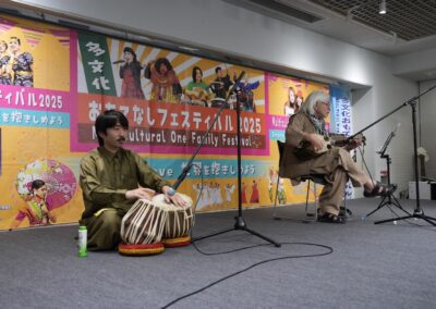 Two musicians perform on stage at the 2025 Multicultural Festival; one plays tabla drums and the other a string instrument, with colorful festival banners in the background.