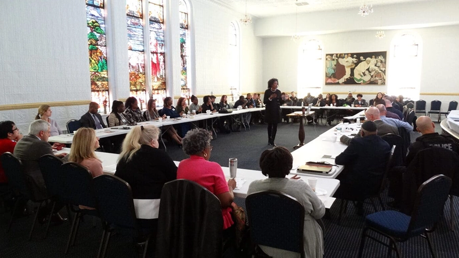 A woman speaks to a large group of people seated around a U-shaped table in a conference room with stained glass windows and a large painting on the wall.