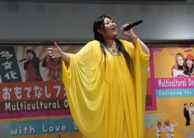 A woman in a flowing yellow dress sings on stage at the One Family Festival 2025, with colorful event banners bringing the multicultural festival to life in the background.