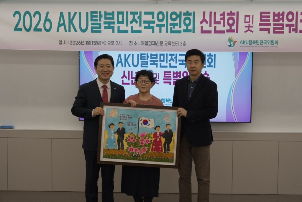 Three people stand indoors holding a framed artwork, with a banner in Korean behind them about a 2026 AKU 탈북민위원회 event supporting North Korean escapees and the Korean Dream citizen movement.