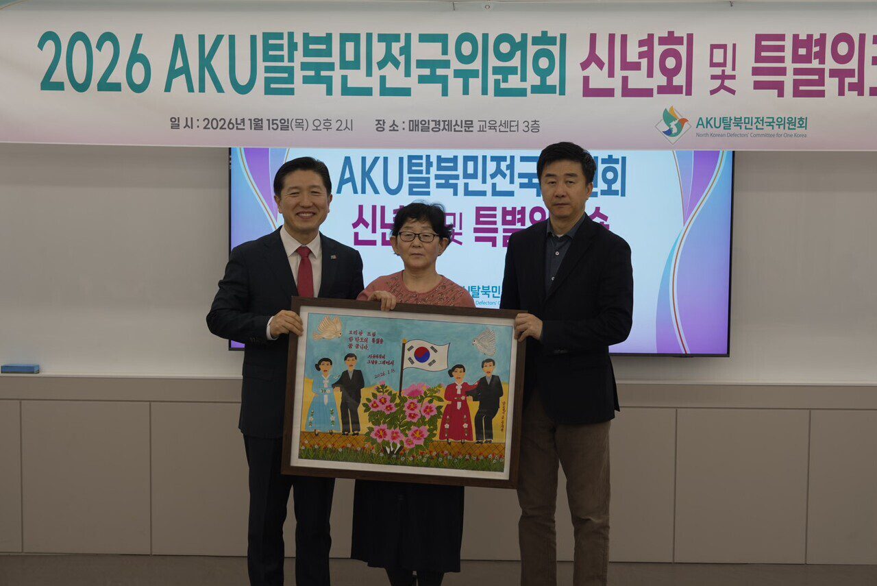 Three people stand indoors holding a framed artwork, with a banner in Korean behind them about a 2026 AKU 탈북민위원회 event supporting North Korean escapees and the Korean Dream citizen movement.