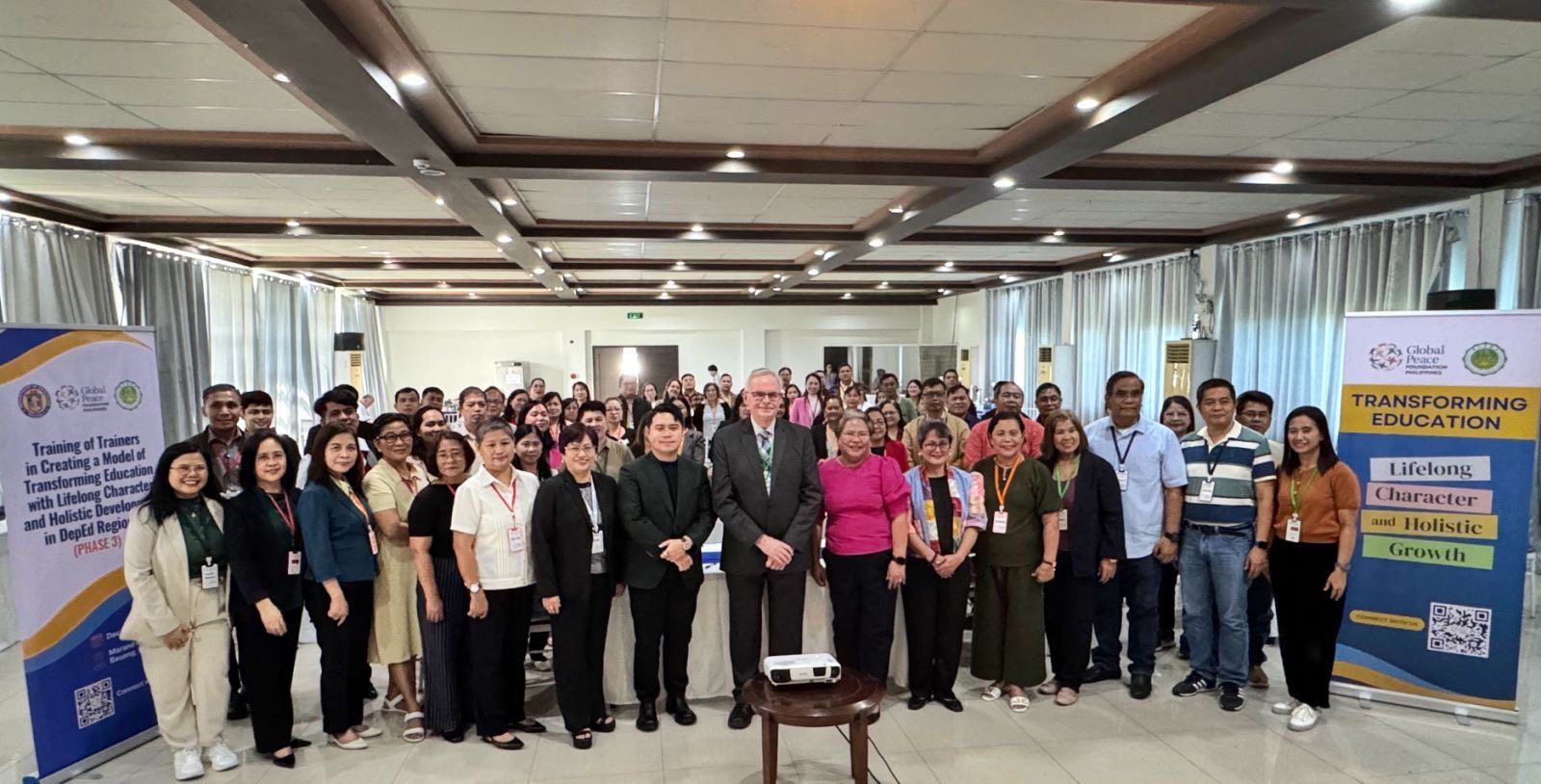 A large group of people pose for a photo in a conference room, flanked by banners about transforming education and holistic leadership for lifelong growth.