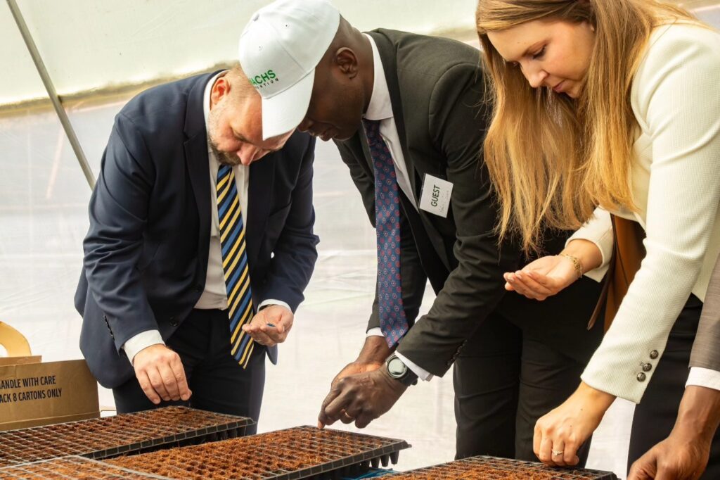 Three people in business attire plant seeds in soil trays inside a greenhouse at the SMACHS Centre of Excellence, participating in a hands-on agricultural activity aligned with the 2025-2030 Strategic Plan.