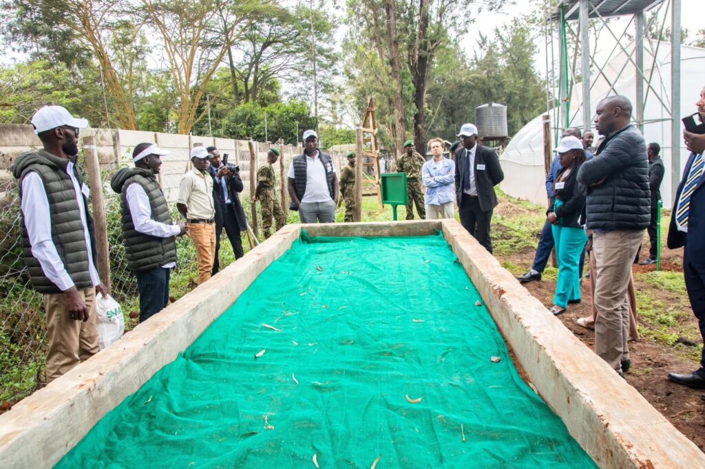 A group of people stands around a rectangular wooden structure covered with green fabric outdoors at the SMACHS Centre of Excellence, with trees and water tanks visible in the background, highlighting GPF Africa's 2025-2030 Strategic Plan.