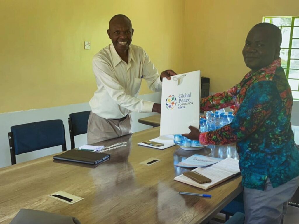 Two men stand beside a conference table, exchanging a gift bag labeled “Global Peace Foundation,” highlighting service initiatives. Notebooks, papers, and bottled water are on the table.