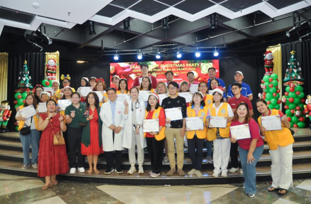 A group of people pose on a stage holding certificates for the Philippines Heal a Heart women project, with Christmas decorations and trees in the background.