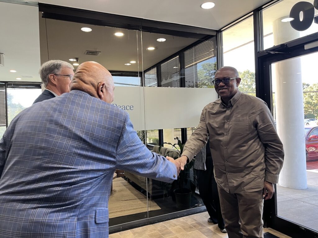 Three men greet each other in an office lobby, with two men shaking hands while another stands nearby—an atmosphere reminiscent of a Youth Assembly featured in Nigeria News.
