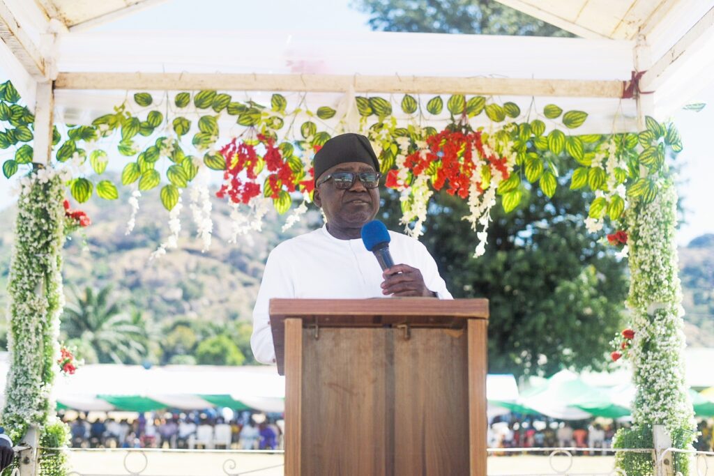 A man in a white shirt and black hat speaks into a microphone at a wooden podium decorated with greenery and flowers, addressing the crowd about community-driven peacebuilding in Nigeria, with trees in the background.