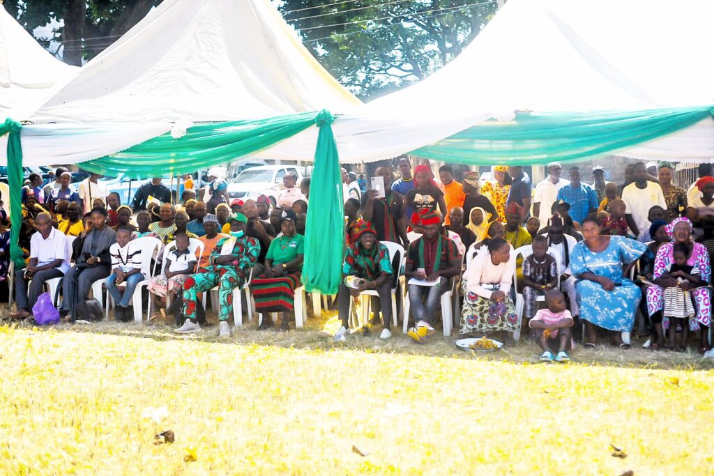 A group of people sit under a large tent with green and white drapes during a daytime outdoor event in Nigeria, highlighting community-driven peacebuilding and a strong sense of cultural identity.