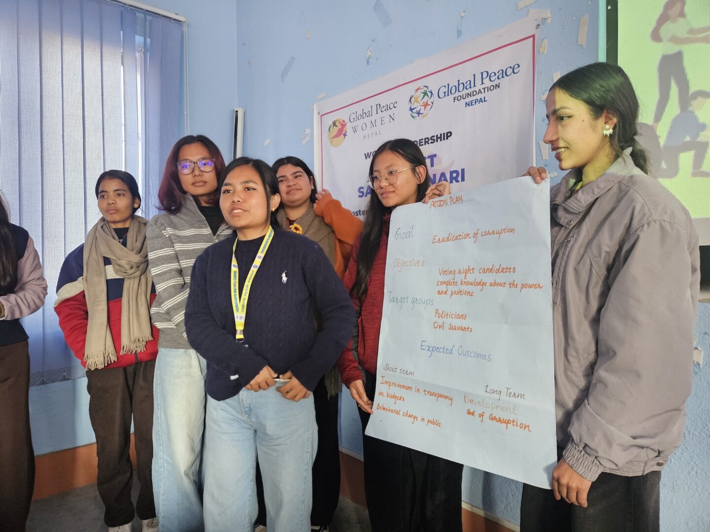 A group of service-minded women stands indoors, one holding a poster with handwritten notes, during a Global Peace Foundation event.
