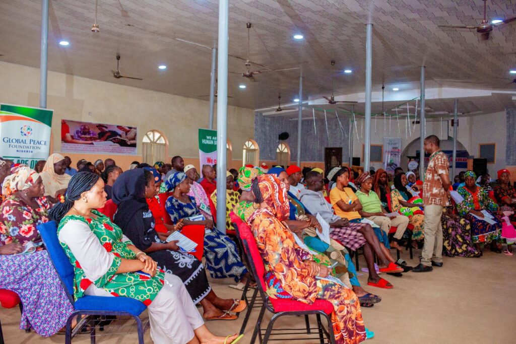 A group of people sits in colorful chairs, listening to a standing speaker in a large, well-lit hall with banners on the walls, as GPF Nigeria discusses the Path to Peace.