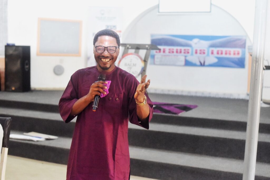 A man in a purple outfit holds a microphone and smiles while speaking on stage in a church, with a