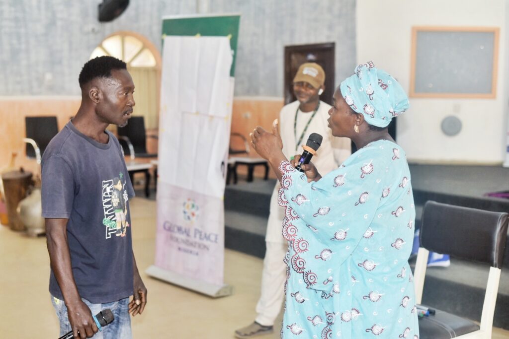A woman holding a microphone speaks to a man in a casual t-shirt as another man stands in the background, engaging in dialogue about Building Peace in a room with chairs and a banner.