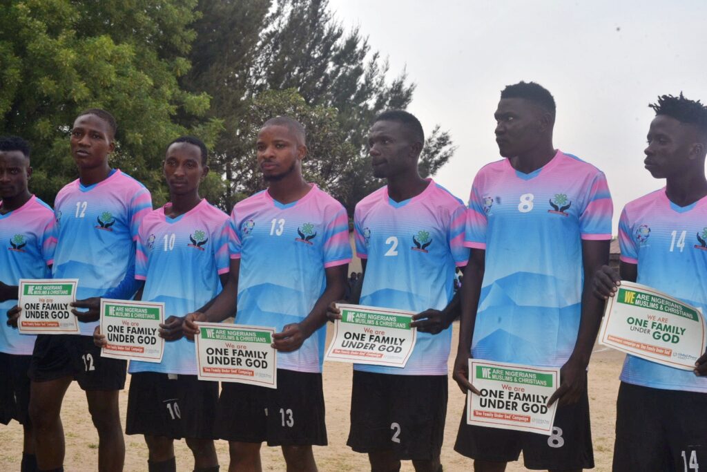 A group of seven male soccer players in matching pink and blue jerseys, representing Hayin Banki, stand outdoors in a line holding signs that read