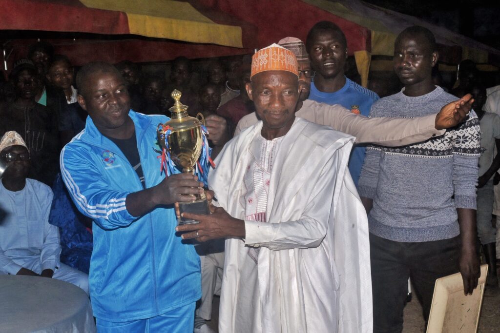 Two men hold a trophy together during an outdoor sports event at night in Kaduna State, surrounded by onlookers under a tent, celebrating teamwork and peace.