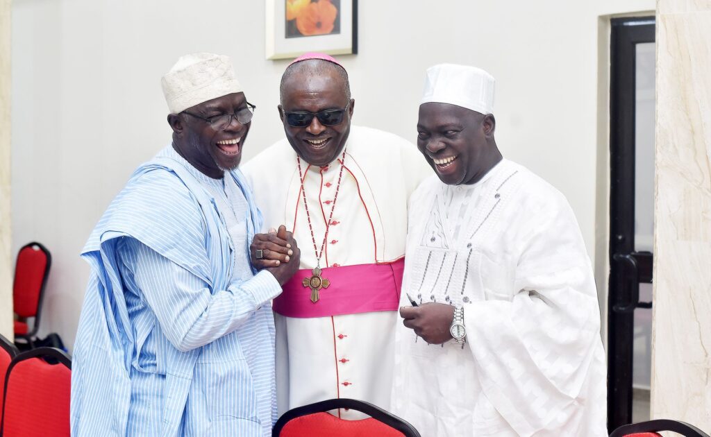 Three men in traditional and clerical attire, likely interfaith leaders, stand together indoors, smiling and holding hands, with red chairs and a framed picture in the background—a gesture promoting lasting peace.