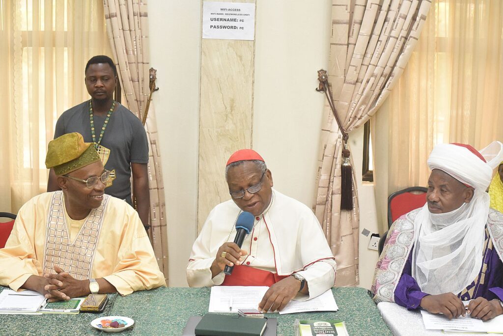 Three interfaith leaders in traditional and religious attire sit at a table during a meeting; the man in the center speaks into a microphone.