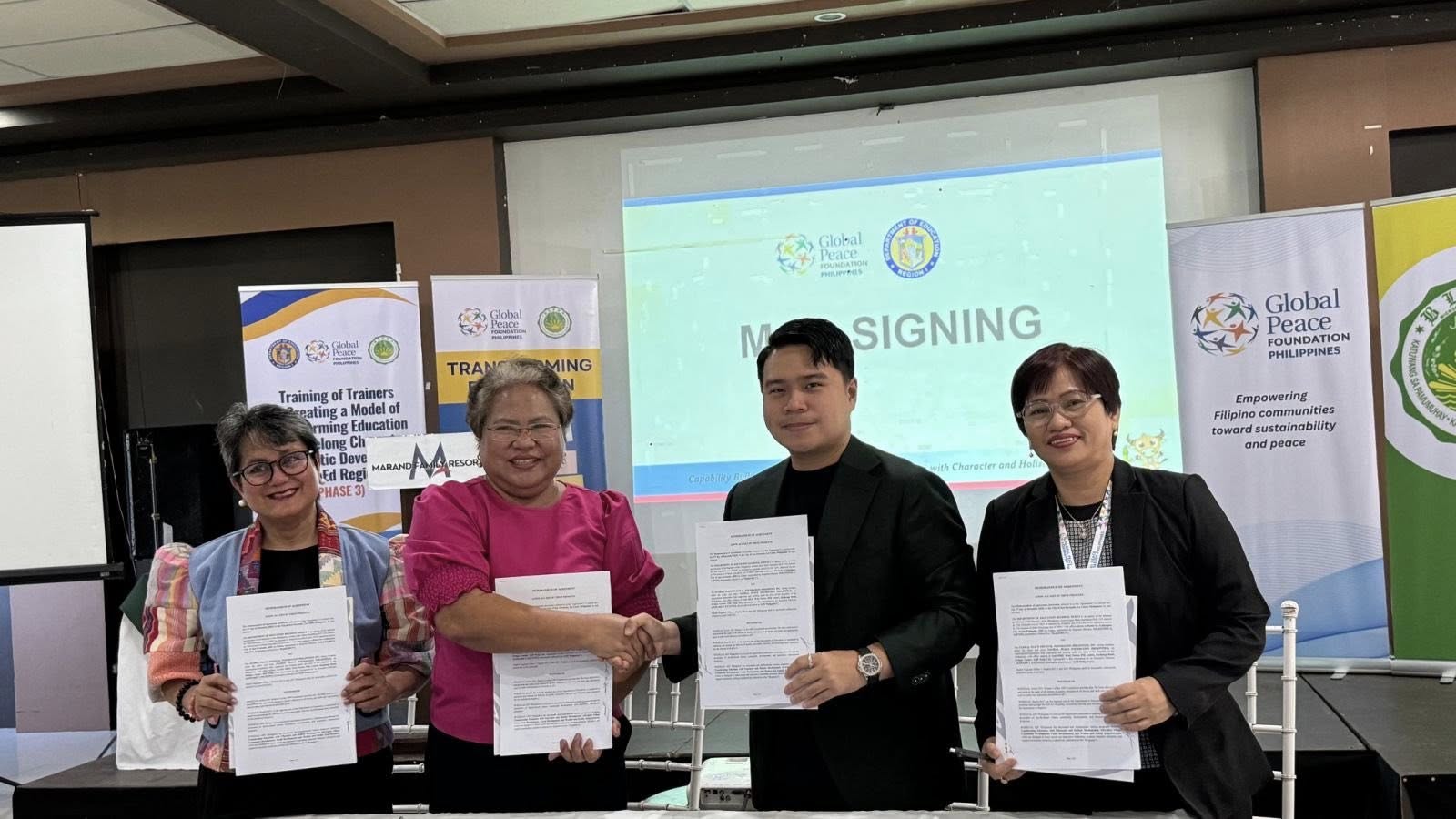 Four people stand side by side holding signed documents in front of a screen displaying