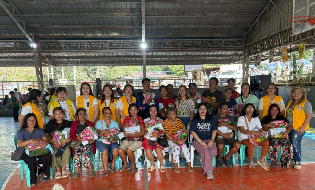 A group of people, some wearing yellow vests, pose together at a covered open-air venue; several seated individuals hold food or gift packages during the Philippines Heal a Heart women project, Dec. 2025.